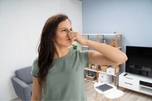 Woman in light green t-shirt holding her hand over her nose and making a face like she smells something bad in the house.